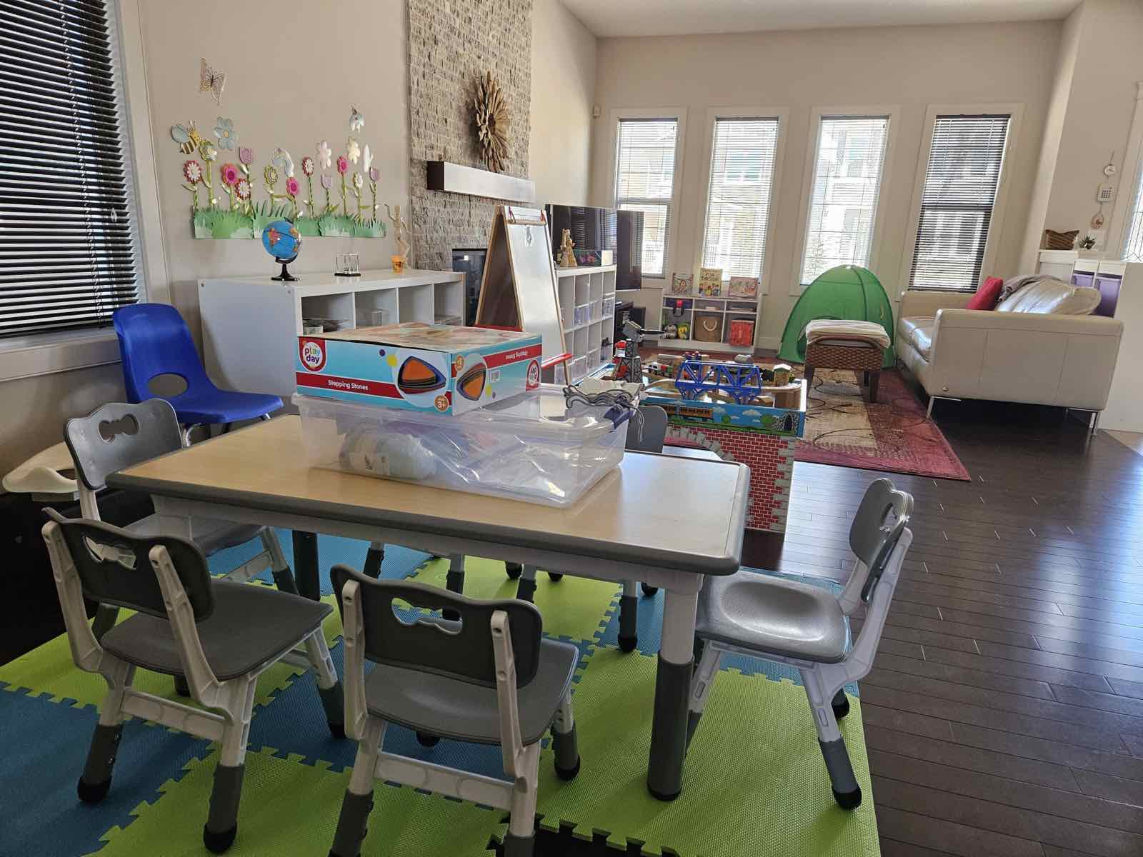 Activity table with kid-sized chairs on a soft play mat, large windows with natural light, train table and reading tent in background